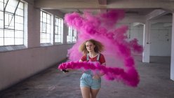 Frontansicht einer jungen kaukasischen Frau mit lockigem Haar, die in einer leeren Lagerhalle eine Rauchmaschine hält, die einen rosafarbenen Rauch produziert — Stockfoto