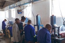Vista lateral de un grupo diverso de hombres jóvenes que trabajan en una fábrica de bolas de cricket hablando juntos mientras operan maquinaria en la línea de producción . - foto de stock