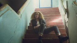 Vue à angle bas d'une jeune femme caucasienne aux cheveux bouclés portant une veste en cuir assise dans les escaliers tout en regardant attentivement la caméra — Photo de stock