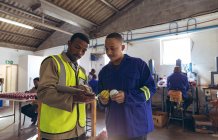 Vista frontal de un joven gerente afroamericano sosteniendo una tableta y hablando con un joven trabajador masculino de raza mixta en el taller en una fábrica de bolas de cricket . - foto de stock