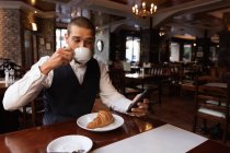 Vista frontal de cerca de un joven caucásico usando su teléfono inteligente y tomando café sentado en una mesa dentro de un café. Nómada digital en movimiento . - foto de stock