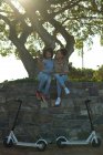 Vue de face de deux jeunes sœurs souriantes de race mixte adultes assises sur un mur dans un parc urbain, utilisant un smartphone et prenant des selfies, leurs scooters électriques garés en dessous d'eux, rétro-éclairés — Photo de stock