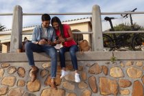 Vista frontale di una giovane coppia mista sorridente seduta fuori su un muro al sole a guardare i loro smartphone — Foto stock