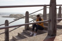 Вид збоку усміхненої молодої змішаної жінки, що сидить на кроках у навушниках і використовує свій смартфон, з пляжем і морем на задньому плані — стокове фото