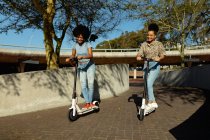 Вид спереду на двох молодих дорослих змішаних сестер, які катаються на електричних скутерах у міському парку, посміхаючись — стокове фото