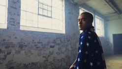 Seitenansicht eines jungen hispanisch-amerikanischen Mannes mit einer amerikanischen Flagge über den Schultern, der aufmerksam in die Kamera blickt, während er in einer leeren Lagerhalle steht — Stockfoto