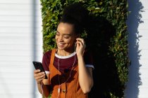 Vista frontal close-up de uma jovem mulher de raça mista sorridente usando seu smartphone com fones de ouvido, de pé ao ar livre ao sol — Fotografia de Stock