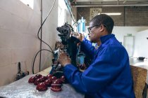 Афроамериканець середнього віку, одягнений в окуляри, працює над вишиванням зовнішніх половинок крикетних кульок, використовуючи швейну машинку на фабриці спортивного устаткування.. — стокове фото