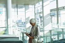 Seitenansicht einer Geschäftsfrau im Hijab, die an einem digitalen Tablet in der Nähe eines Geländes in einem modernen Büro arbeitet. — Stockfoto