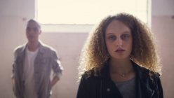 Front view of a Caucasian woman with curly hair and wearing a leather jacket looking intently at the camera while a young man stands in the background inside an empty warehouse — Stock Photo