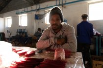 Porträt eines jungen afrikanisch-amerikanischen Mannes, der an einer Werkbank in einer Fabrik sitzt und Cricketbälle herstellt, in die Kamera blickt und lächelt. — Stockfoto