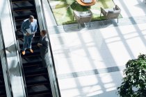 Vista de ángulo alto de hombres de negocios que interactúan entre sí mientras se mueven abajo en escaleras mecánicas en la oficina . - foto de stock