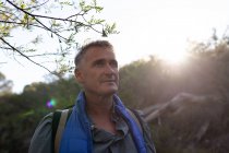 Front view close up of a mature Caucasian man wearing a backpack looking up at the scenery during a hike, backlit by sunlight — Stock Photo