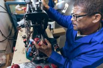 Elevated side view of a middle aged African American man wearing glasses working on the stitching of the shaped outer halves of cricket balls using a sewing machine in a workshop at a sports equipment factory. — Stock Photo