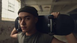 Close up view of a young Hispanic-American man with piercings wearing a dark grey shirt and beanie while holding a skateboard over shoulder and looking intently at the camera while standing inside an empty warehouse — Stock Photo