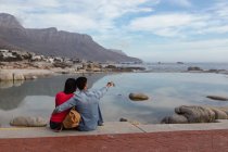 Vista posteriore di una giovane coppia mista seduta fuori su un muro che abbraccia e guarda verso il mare, l'uomo indica la distanza — Foto stock