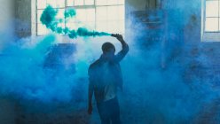 Frontansicht eines jungen hispanisch-amerikanischen Mannes, der eine graue Jacke über einem weißen Hemd trägt und eine Rauchmaschine in der Hand hält, die blauen Rauch innerhalb einer leeren Lagerhalle produziert — Stockfoto