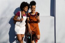 Vista frontal de dos hermanas adultas jóvenes de raza mixta, una llevando una mochila y la otra sosteniendo un monopatín, sonriendo y mirando a un teléfono inteligente, apoyado contra una pared al sol - foto de stock