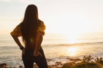 Nahaufnahme einer jungen Mischlingsfrau mit langen Haaren, die am Strand steht, die Hände in den hinteren Taschen ihrer Jeans und bei Sonnenuntergang aufs Meer blickt — Stockfoto