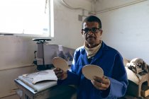 Retrato de um homem afro-americano de meia-idade usando óculos trabalhando em uma fábrica fazendo bolas de críquete, olhando para a câmera e segurando couro cortar formas . — Fotografia de Stock