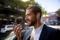 Vue de côté gros plan d'un jeune homme caucasien souriant parlant sur un smartphone le tenant devant son visage et portant des écouteurs dans une rue de la ville. Nomade numérique en mouvement . — Photo de stock