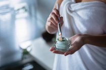 Mid section of a woman wearing a bath towel holding a jar of face pack and dipping in a brush in preparation to apply it to her face — Stock Photo