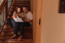 Передній погляд на чоловіка і жінку середнього віку, які сидять на сходах у своєму домі і користуються комп 