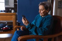 Вид збоку старший кавказька жінка сидить у кріслі, використовуючи смартфон на дому — стокове фото