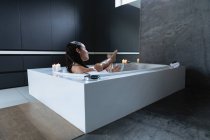 Vue latérale d'une jeune femme brune caucasienne assise dans un bain en mousse avec des bougies allumées sur le côté, les pieds levés, dans une salle de bains moderne — Photo de stock