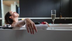Vista lateral de uma jovem mulher morena caucasiana deitada em um banho com uma vela acesa e uma taça de champanhe ao lado, inclinando-se para trás com os olhos fechados — Fotografia de Stock