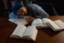 Vista frontal close-up de uma jovem estudante asiática vestindo um turbante dormindo em uma mesa cercada por livros durante o estudo em uma biblioteca — Fotografia de Stock