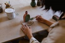 Sur l'épaule d'une potière assise à une table de travail peignant une glaçure colorée sur une fiole d'argile dans un atelier de poterie — Photo de stock