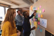 Side view of a young Caucasian woman and man standing and watching their young African American male colleague writing on an ideas board during a team brainstorm session at a creative office — Stock Photo