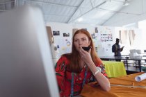 Frontansicht einer jungen kaukasischen Modestudentin, die ein Smartphone in der Hand hält und spricht und einen Computer in einem Atelier der Modehochschule benutzt, während eine Studentin im Hintergrund arbeitet — Stockfoto