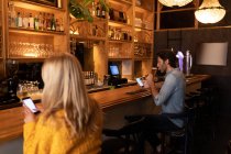 Vista lateral de un joven hombre y una mujer caucásicos relajándose de vacaciones en un bar, bebiendo vino y cerveza usando teléfonos inteligentes - foto de stock
