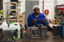 Vista frontal de perto de um jovem trabalhador da fábrica afro-americano sentado e inspecionando equipamentos na loja de máquinas em uma fábrica de processamento, com prateleiras de equipamentos em segundo plano — Fotografia de Stock