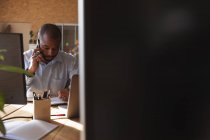 Nahaufnahme eines jungen afrikanisch-amerikanischen Mannes, der in einem kreativen Büro an einem Schreibtisch sitzt und mit einem Smartphone spricht, gesehen zwischen Computerbildschirmen — Stockfoto