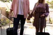 Vue de face au milieu d'un couple se relaxant en vacances debout avec des valises à l'extérieur d'un hôtel, se tenant la main — Photo de stock