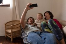 Вид молодых кавказских родителей, лежащих на диване со своим ребенком и делающих селфи — стоковое фото