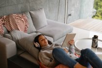 Vista elevada de uma jovem morena caucasiana reclinada em um sofá com as pernas para cima, usando fones de ouvido e assistindo a um tablet — Fotografia de Stock