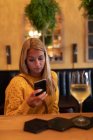 Вид на молоду Кавказьку жінку розслабляє на відпочинку в барі, випиваючи вино і за допомогою смартфона — стокове фото