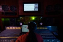 Rear view close up of a young mixed race male sound engineer sitting and working at a mixing desk in a recording studio wearing headphones — Stock Photo