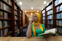 Передній погляд на молоду азіатську студентку, одягнену в хіджаб, яка тримає книжку і навчається в бібліотеці. — стокове фото