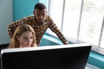 Vista frontal de perto de um jovem afro-americano de pé e uma jovem caucasiana sentados em uma mesa juntos, olhando para um monitor de computador no escritório moderno de um negócio criativo — Fotografia de Stock