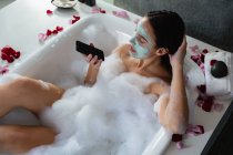 Vista elevada de uma jovem caucasiana vestindo um pacote facial, sentada em um banho de espuma com pétalas de rosa ao redor da borda usando um smartphone — Fotografia de Stock