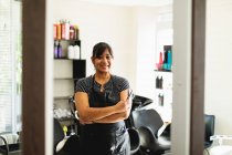 Портрет женщины-парикмахера средних лет, улыбающейся в камеру со скрещенными руками в парикмахерской — стоковое фото