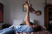 Seitenansicht eines jungen kaukasischen Vaters, der sein Baby über sich hält und auf einem Bett liegt — Stockfoto