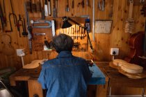 Visão traseira de uma luthier feminina caucasiana sênior trabalhando em sua oficina, com ferramentas penduradas na parede no fundo — Fotografia de Stock