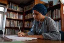 Vista lateral de cerca de una joven estudiante asiática con un turbante escribiendo y estudiando en una biblioteca - foto de stock