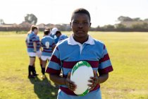 Porträt einer jungen erwachsenen afrikanisch-amerikanischen Rugbyspielerin, die auf einem Rugbyfeld steht und einen Rugbyball in die Kamera hält, während ihre Teamkolleginnen im Hintergrund miteinander reden — Stockfoto